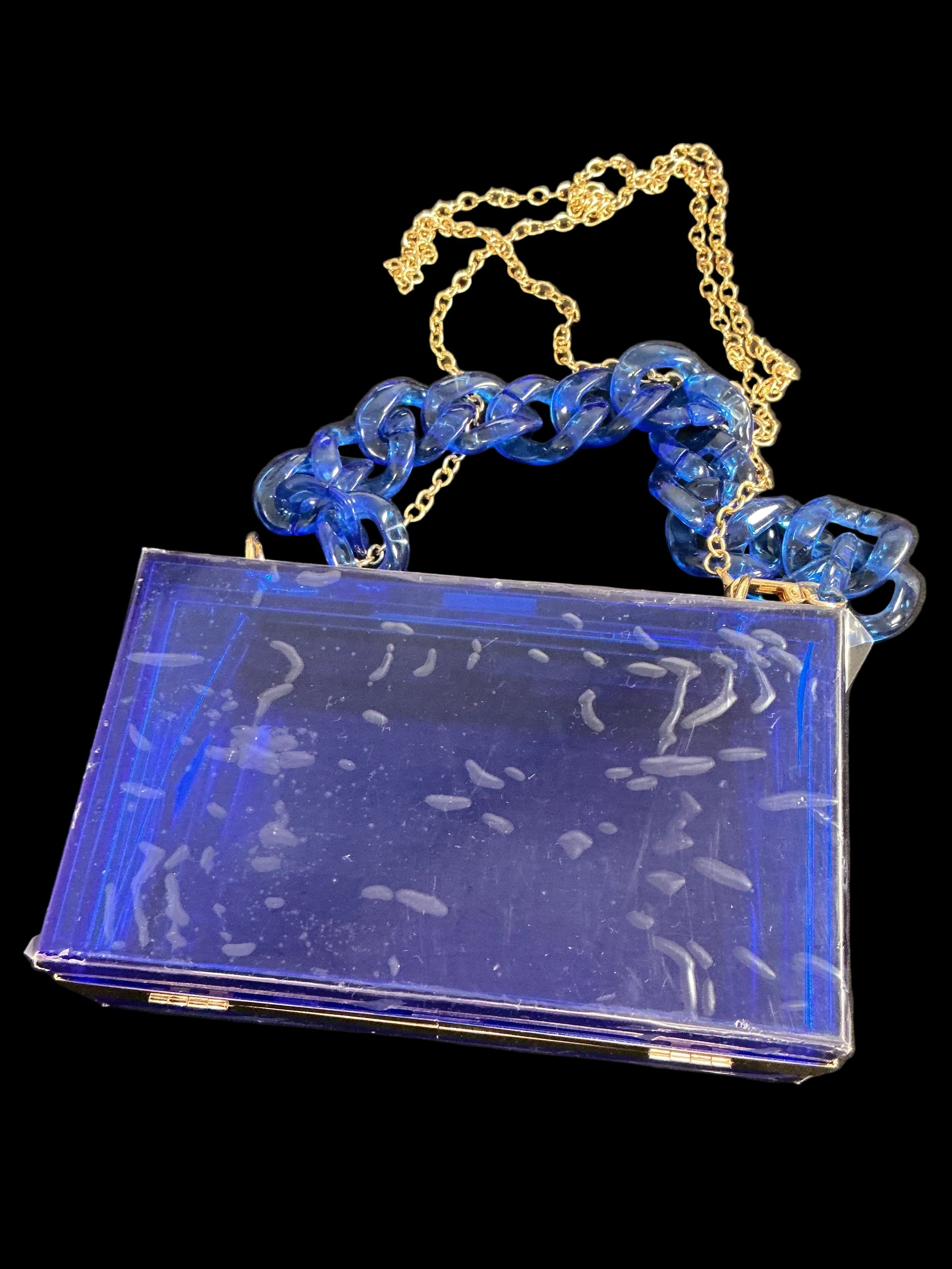 A vintage-style rectangular clutch bag with a translucent cobalt blue body, a chunky blue chain handle, and a gold-tone chain shoulder strap, resting on a dark surface.